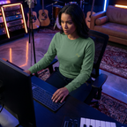 Person in a green shirt sitting at a desk with electronic equipment in a room with musical instruments.