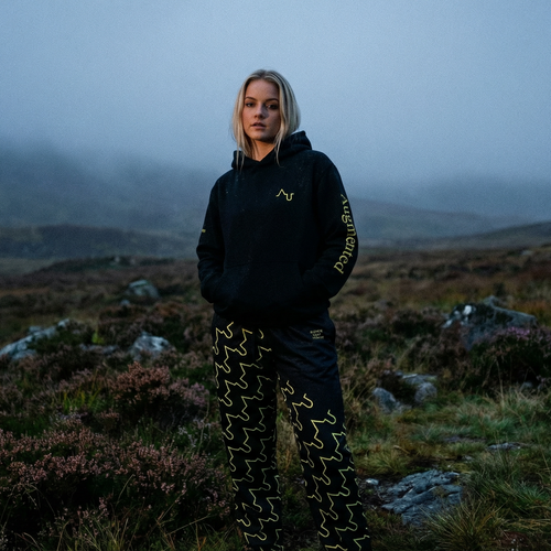 Person standing in a misty, mountainous landscape wearing a black jacket with a logo.