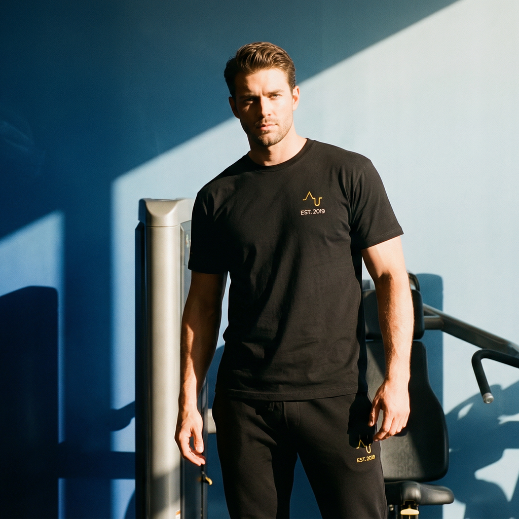 Man in black t-shirt and pants standing in a gym with blue wall and equipment.