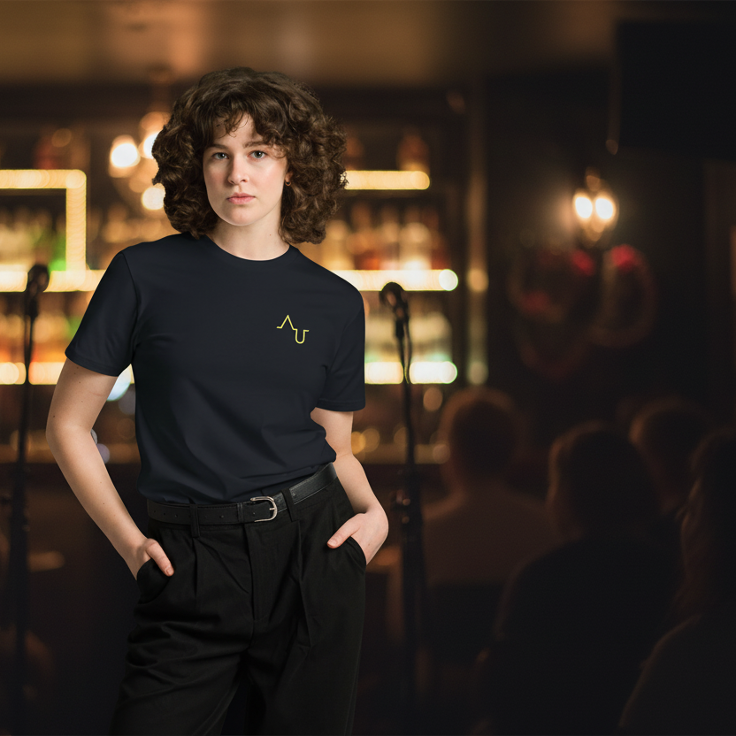 Person wearing a black t-shirt with a logo in a dimly lit room with blurred lights and people in the background