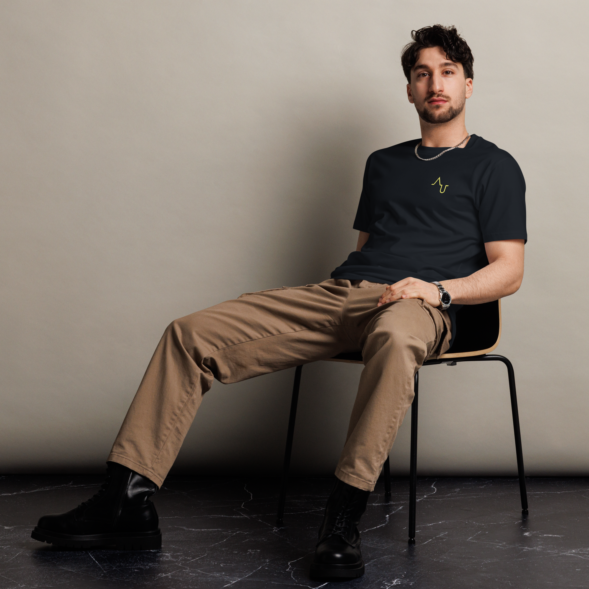 Man sitting on a chair wearing a black t-shirt and beige pants against a plain background